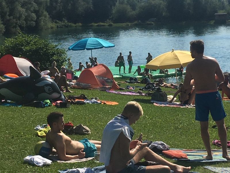 Menschen genießen einen sonnigen Tag in einem Park am Seeufer mit Zelten, Sonnenschirmen und Schlauchbooten.