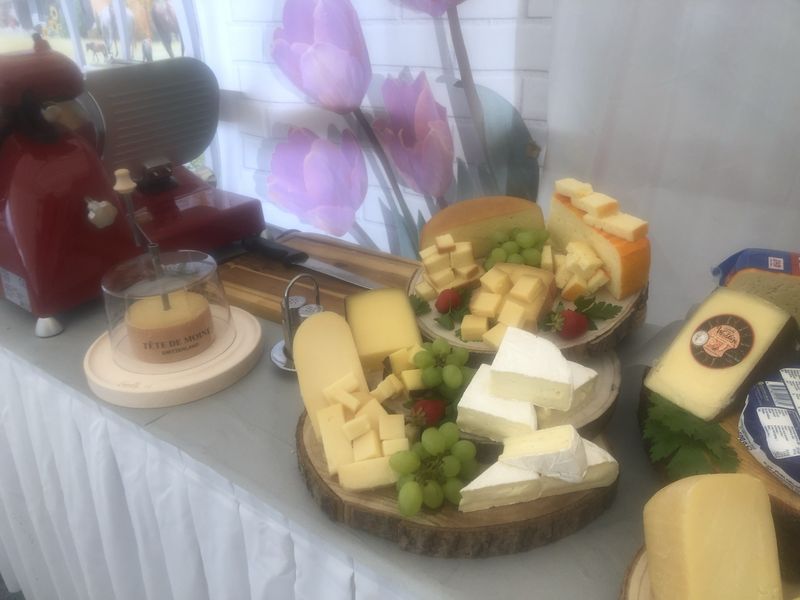 Verschiedene Käsesorten auf einem Holzbrett, im Hintergrund Trauben, ein rotierender Käsehobel und Tulpen.