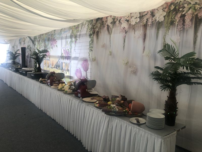 Ein langer Buffettisch, dekoriert mit Blumenarrangements und verschiedenen Gerichten, aufgebaut in einem Zelt mit weißen Tischdecken.